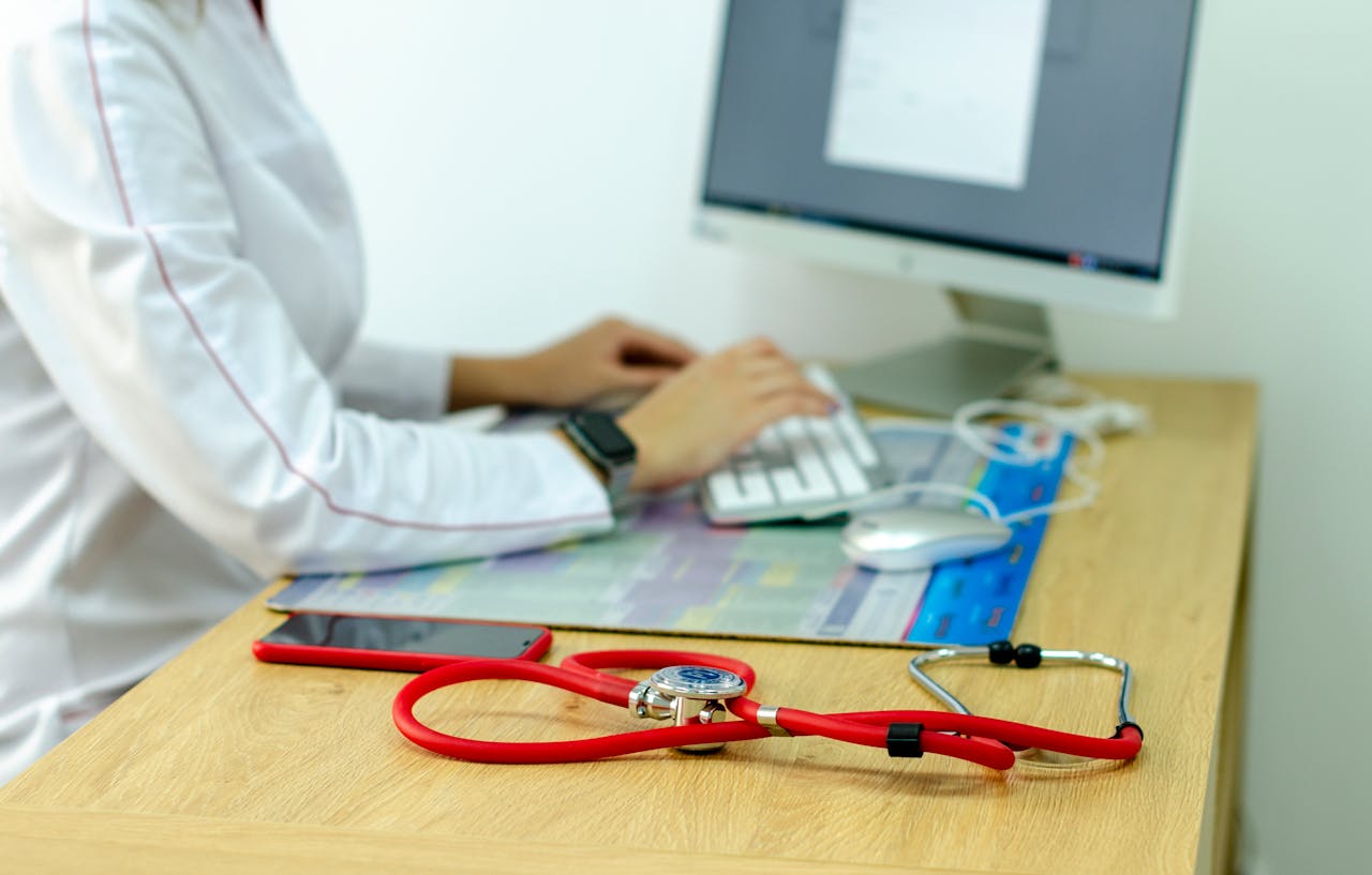 A medical professional typing on a computer with a stethoscope on a wooden desk, indicating a modern healthcare setting.